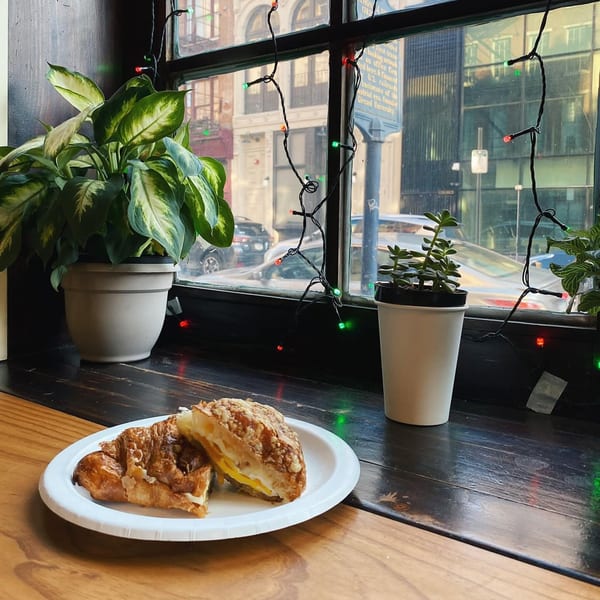 A croissant sandwich on a white plate sits on a wooden counter by a window with string lights.