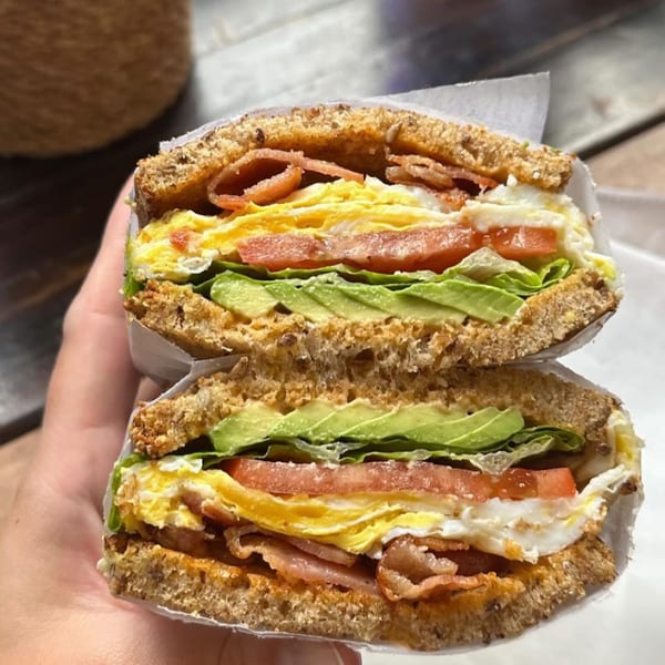 A hand holds a hearty breakfast sandwich with bacon, egg, avocado, tomato, and lettuce on whole wheat bread.