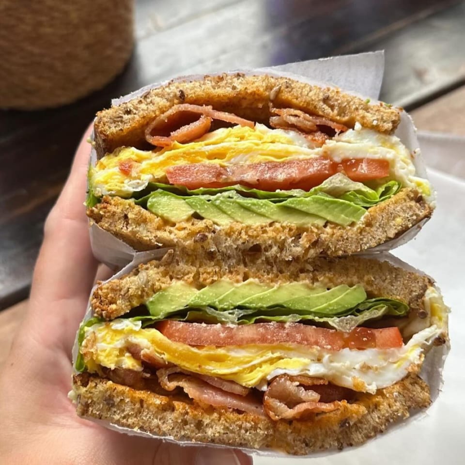 A hand holds a hearty breakfast sandwich with bacon, egg, avocado, tomato, and lettuce on whole wheat bread.