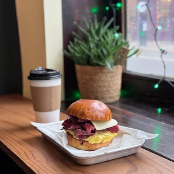 A breakfast sandwich with pastrami, egg, and cheese, next to a coffee cup on a wooden counter.